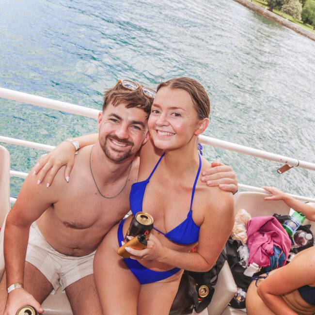 A smiling man and woman in swimwear sit close together on a boat, holding drinks. The woman wears a blue bikini, and the man wears light-colored swim trunks. Water and greenery are visible in the background.