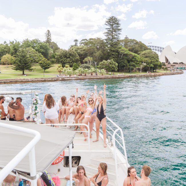 A group of people in swimsuits enjoy a party on a boat in Sydney Harbour, with the Sydney Opera House and Harbour Bridge visible in the background on a sunny day.