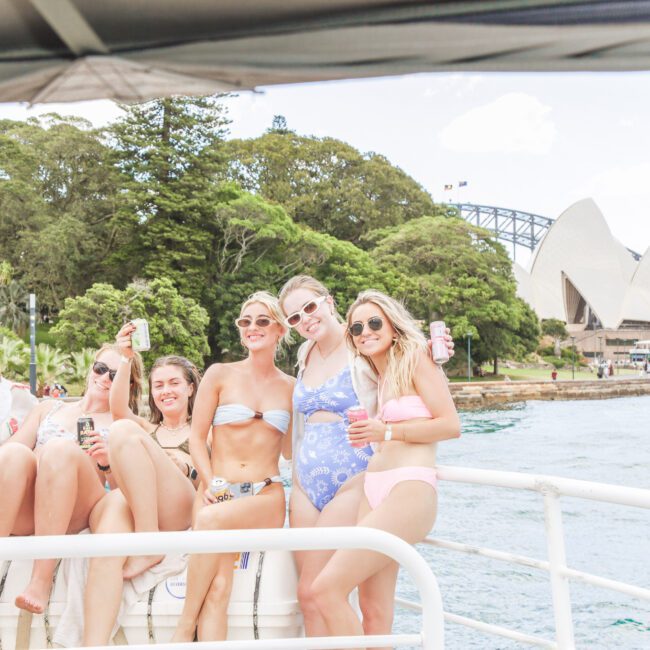 Five women in swimsuits sit and smile on a boat, holding cans, with the Sydney Opera House and lush greenery in the background on a sunny day.