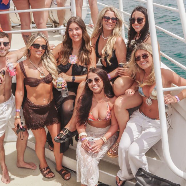 A group of seven women and one man in swimsuits pose and smile together on the deck of a boat, holding drinks, with clear blue water and a life preserver visible in the background.