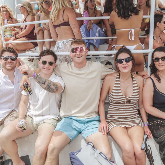 Five young adults sit smiling on a boat with drinks in hand, surrounded by other people in swimwear. The sunny scene suggests a lively party atmosphere on the water.
