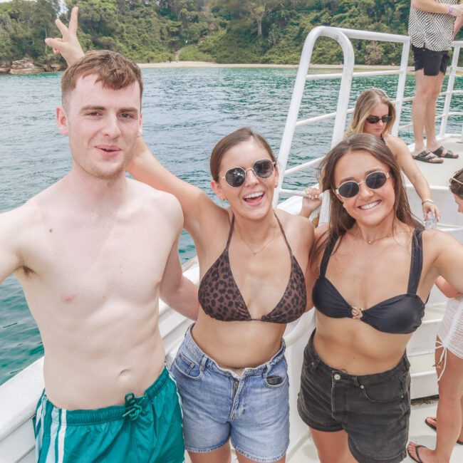Three smiling young adults in swimsuits holding drinks, posing on a boat with people and a lush green coastline in the background, enjoying a sunny day on the water.