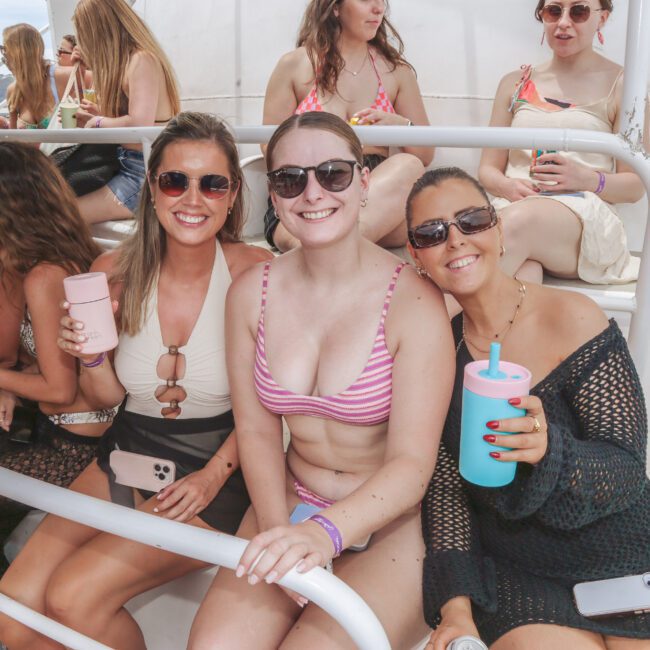 Three women in swimsuits and sunglasses sit closely together on white bleachers, smiling at the camera and holding colorful drink cups. More women in swimsuits are seated and chatting in the background.