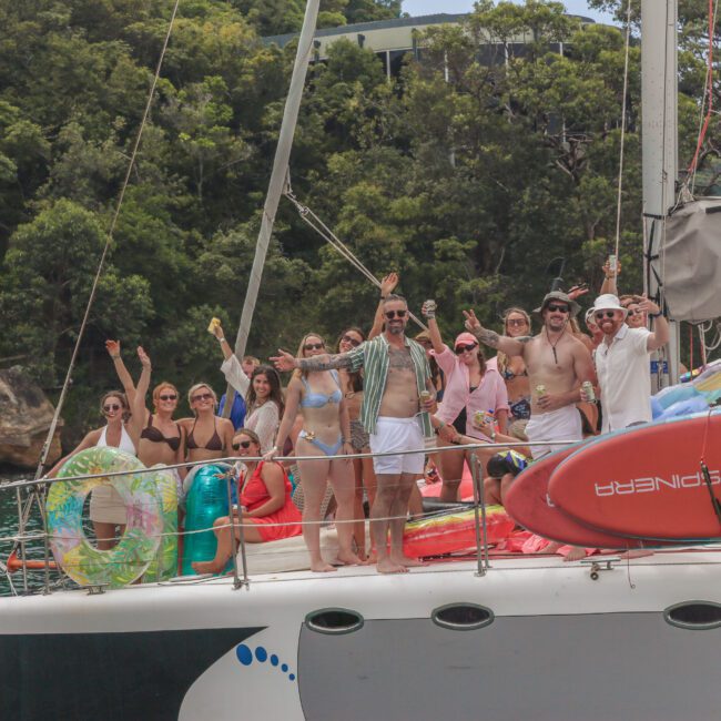 A group of people in swimsuits and summer attire stand on a yacht, smiling and raising their arms. Colorful pool floats and a red paddleboard are visible. Lush green trees and water are in the background.