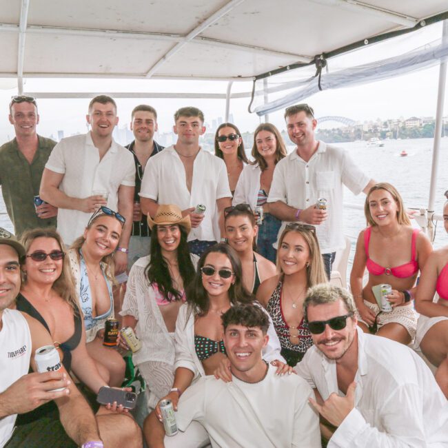 A group of people smiling and posing together on a boat, holding drinks. They are dressed in casual summer clothing, with water and a city skyline visible in the background.