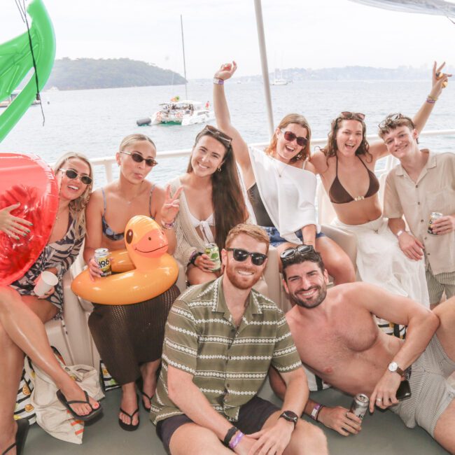 A group of smiling friends on a boat pose for a photo, holding drinks and wearing summer outfits. Large inflatable cherry and duck floaties are in the foreground, with water and boats visible in the background.