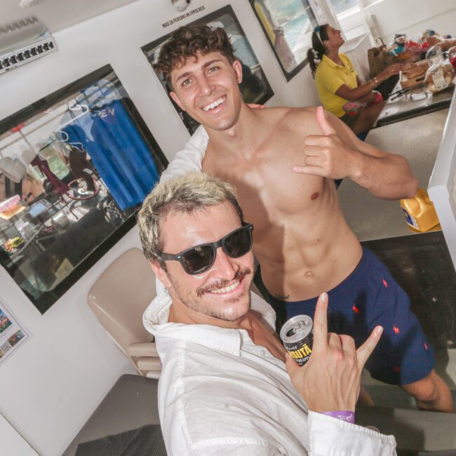 Two smiling men pose on a boat, one shirtless and making a shaka sign, the other wearing sunglasses and holding a drink can. Other people and tables with food are visible in the background.