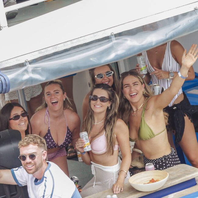 A group of smiling young adults in swimsuits pose and wave while on a boat. Some hold drinks and wear sunglasses. Bottled water and snacks are on the table in front of them. The atmosphere is lively and festive.