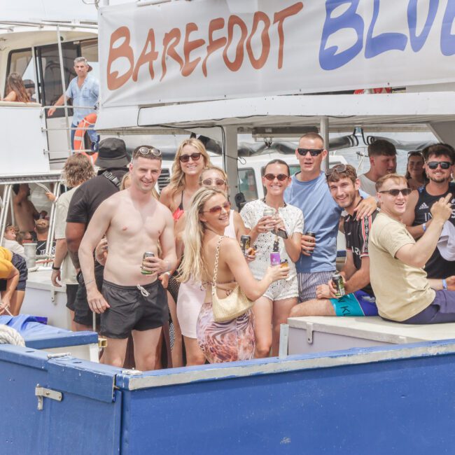 A group of smiling people in summer clothes stand and sit on a boat named "Barefoot Blue," holding drinks and posing for a photo during a lively outdoor gathering on the water.