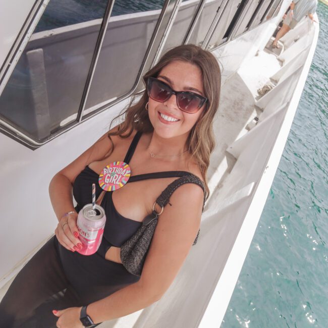 A smiling woman wearing sunglasses and a "Birthday Girl" badge holds a canned drink while standing on the deck of a boat above turquoise water. The sun is shining, and she appears to be enjoying a celebration.