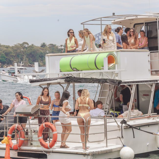 A group of people are socializing on a white yacht, some standing on the deck and others on an upper level, with water and city buildings in the background. The atmosphere appears relaxed and festive.