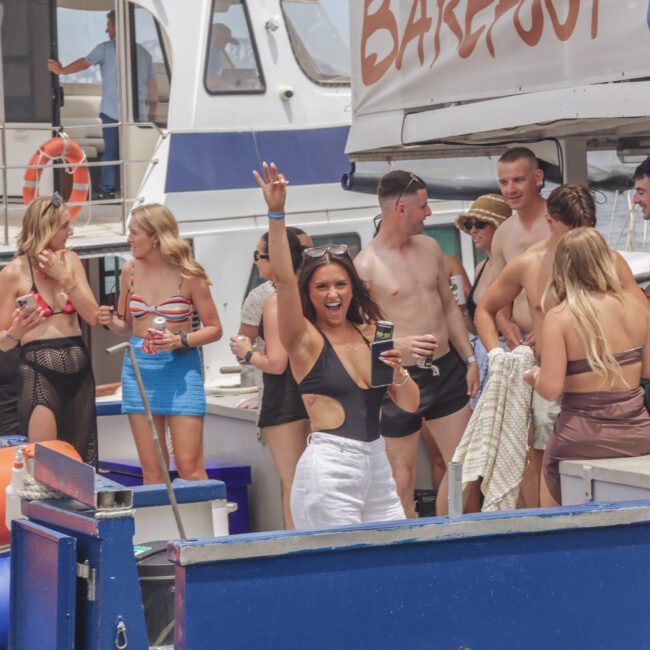A group of young adults in swimsuits enjoy a lively boat party, with some dancing and holding drinks. One woman in the foreground raises her arm and smiles, while others socialize near the edge of the boat.