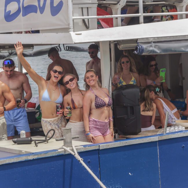 A group of young adults in swimwear smile, pose, and hold drinks on a blue and white boat during a lively daytime party. Some wave at the camera while others socialize in the background.