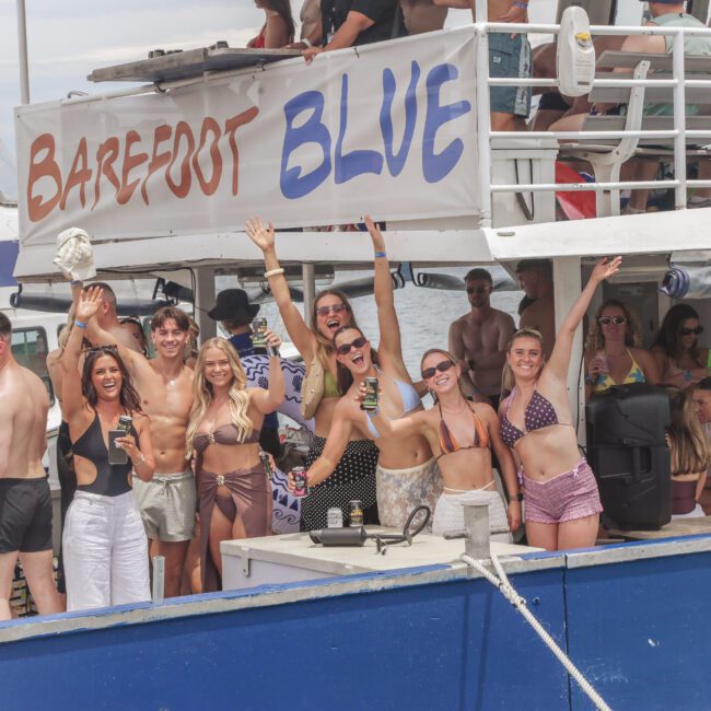 A group of smiling people in swimsuits wave and pose on a crowded boat labeled “Barefoot Blue,” enjoying drinks and music during a lively summer party.
