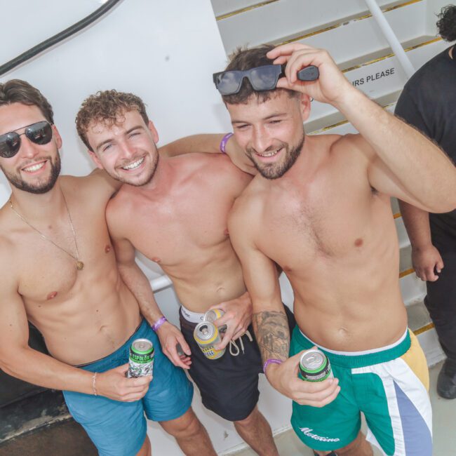 Three young men in swim trunks, smiling and holding canned drinks, pose together on a boat. A security guard stands nearby. The group appears to be enjoying a casual, sunny day at a social event on the water.