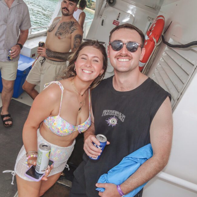 A smiling woman in a floral bikini and a man in sunglasses and a black tank top pose together on a boat with drinks in hand. Other people stand in the background near the water, enjoying the sunny day.