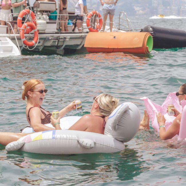 Three women float on inflatable loungers in the water, laughing and drinking canned beverages, with boats and other people in the background on a sunny day.