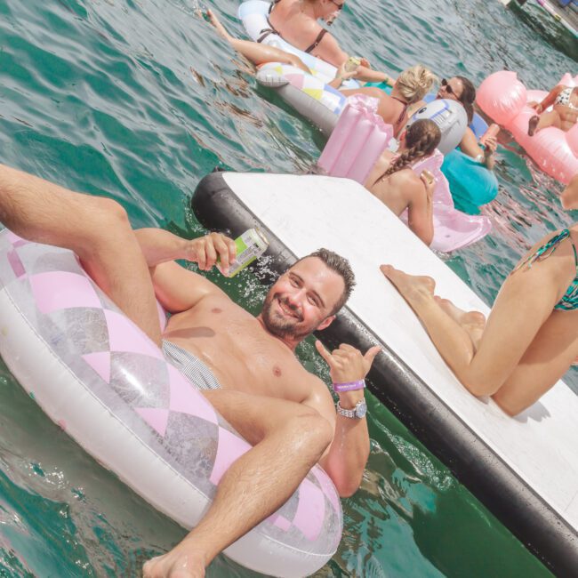 A man relaxes on a pink and white inflatable ring in the water, smiling and holding a drink, with other people on floats and a large floating platform nearby during a sunny boat party.