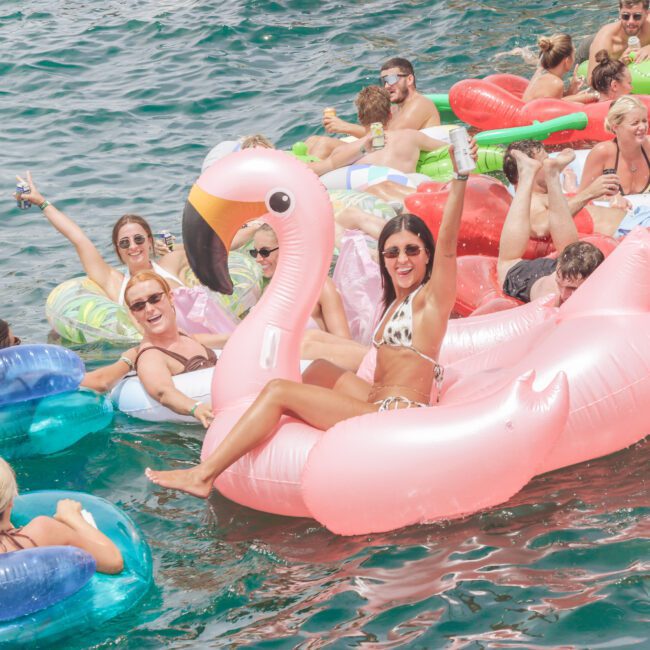 A group of people relax on colorful inflatable floats in the water, including a large pink flamingo float. They are smiling, chatting, and enjoying a sunny day outdoors.