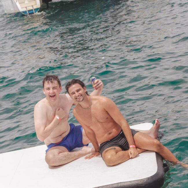 Two young men in swim trunks sit on a floating dock, smiling and posing for the camera. One holds a drink, and both have wet hair, suggesting they were swimming. The water around them is a deep blue-green.
