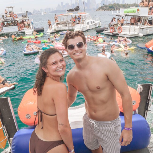 A smiling woman in a brown bikini and a man in swim trunks and sunglasses stand on a boat, with many people on floats and other boats enjoying a lively party on the water in the background.