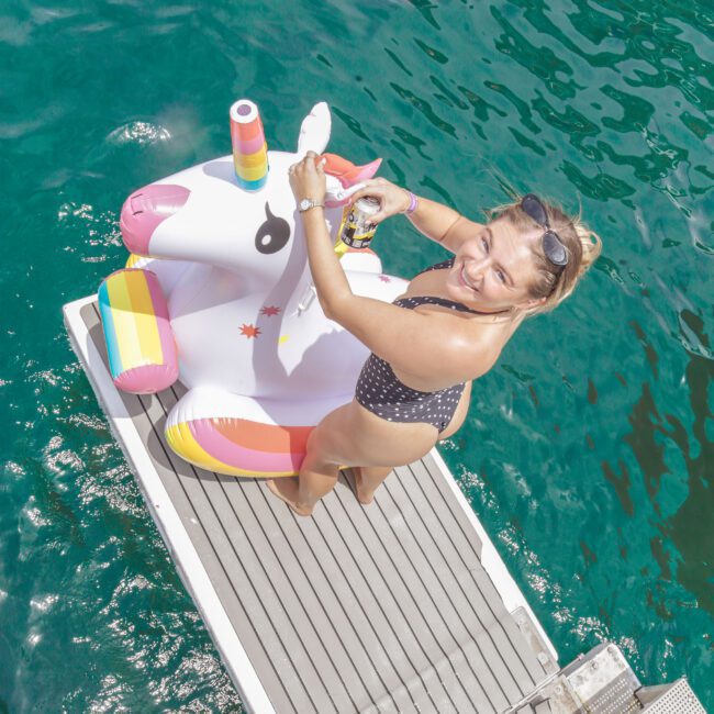 A woman in a polka dot swimsuit smiles while sitting on a unicorn pool float on a dock above clear blue-green water. She holds sunglasses and looks up at the camera in bright sunlight.