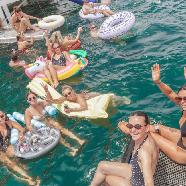 A group of people in swimsuits relax on inflatable pool floats and in the water near a boat, smiling and waving at the camera during a sunny day on the water.