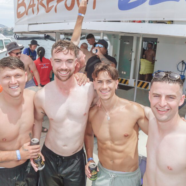 Four smiling young men stand shirtless on a boat, holding drinks, with other people in the background enjoying a sunny day by the water. A boat and "BAREFOOT" sign are visible behind them.