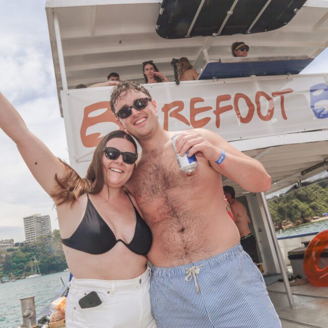 A smiling woman in a black bikini and a man in swim trunks pose together holding drinks on a boat, with a “Barefoot Blue” banner behind them and people in the background enjoying a sunny day by the water.