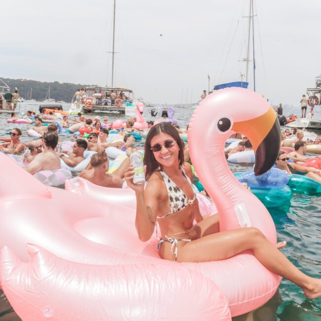A smiling woman in sunglasses and a bikini sits on a large pink flamingo float, holding a drink. She is surrounded by other people on floats in the water, with boats and a hazy sky in the background.
