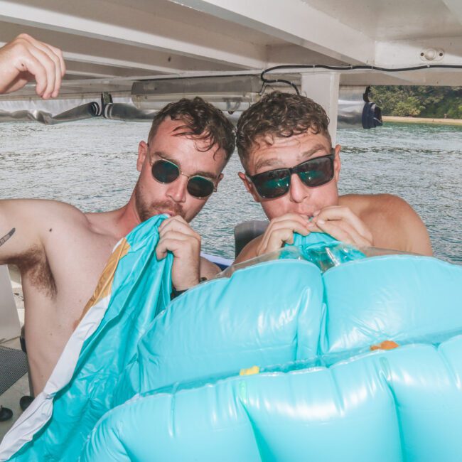 Two men wearing sunglasses are inflating a large blue pool float on a boat, with water and greenery visible in the background. Sunlight filters onto the deck and part of another person is seen at the edge of the frame.