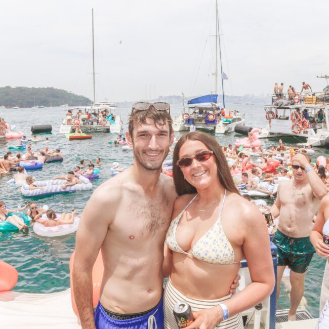 A smiling man and woman in swimsuits pose on a boat at a crowded floating party with many people on inflatables and boats in the background, enjoying a sunny day on the water.