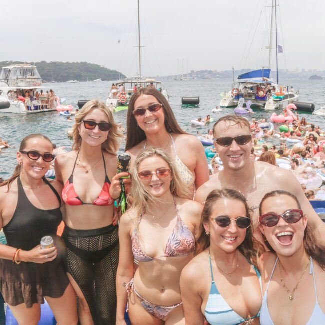 A group of smiling friends in swimwear pose on a boat with a lively crowd of people on floaties and boats in the background during a sunny, festive day on the water.