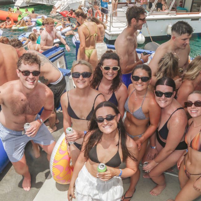 A group of smiling young adults in swimsuits and sunglasses pose on a boat with drinks, surrounded by others swimming and lounging on inflatables in clear blue water under sunny skies.