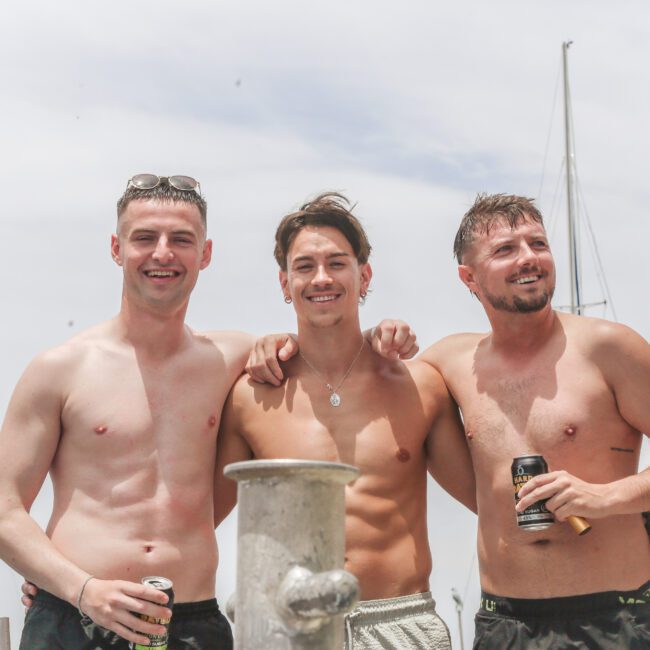 Three young men stand shirtless on a boat, smiling and posing with drinks in hand. Sailboat masts are visible in the background under a cloudy sky.