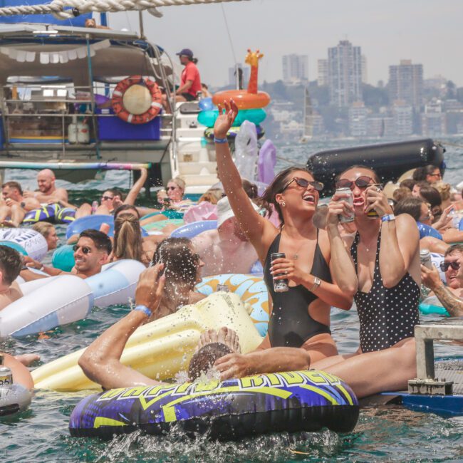 A large group of people in swimsuits relax on colorful inflatables in the water near docked boats, enjoying a lively pool party under the sun with city buildings visible in the background.