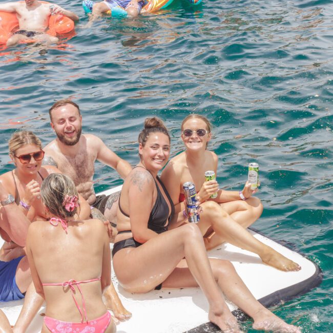 A group of smiling people in swimsuits sit on a floating mat in bright blue water, holding drinks and enjoying a sunny day. Other swimmers and floaties are visible in the background.