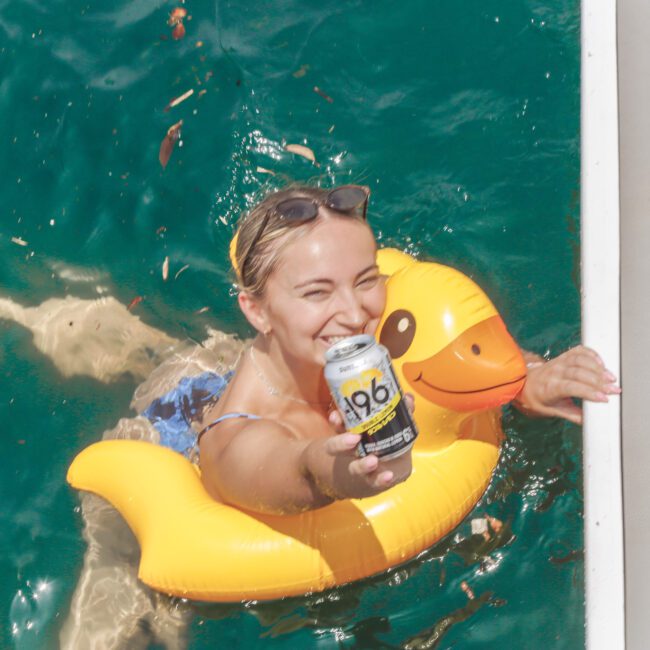 A smiling woman in a pool floats on a yellow duck inflatable and holds up a can of 196 Double Lemon drink toward the camera near a dock.