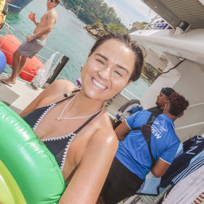 A smiling woman in a swimsuit holds a green inflatable pool float on a sunny boat deck, surrounded by other people, with water and city buildings in the background.