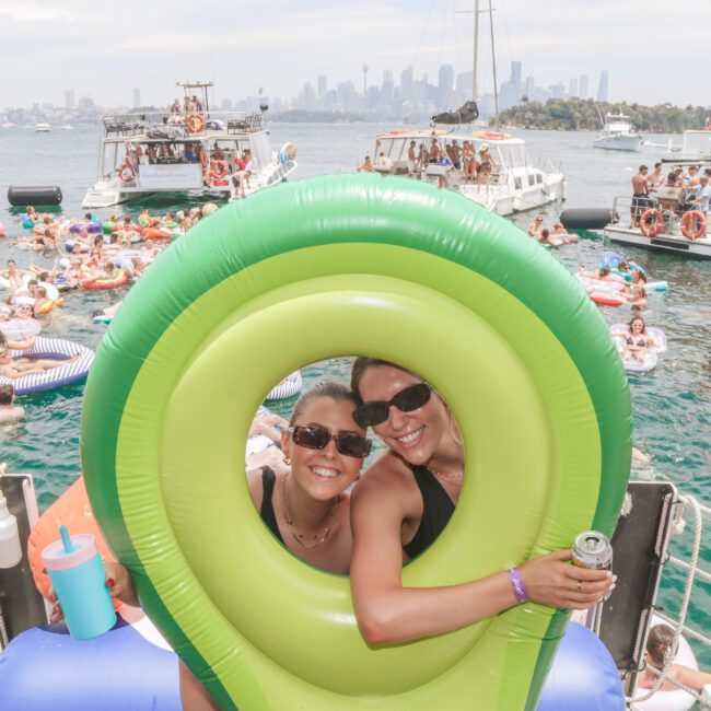 Two people smile through a hole in a green pool float on a crowded boat. Many others float in the water on inflatables, with boats and a city skyline visible in the background. It’s a lively summer scene.