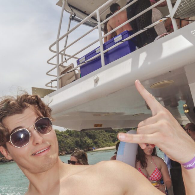 A shirtless man wearing sunglasses poses with a hand gesture on a boat, while a woman stands on the upper deck holding a drink. Other people relax in the background, and the boat is docked near a green, tree-covered shore.