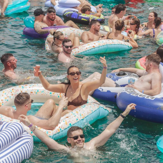 A crowd of people relax and have fun on colorful inflatable rafts and floats in a sunny body of water, smiling, laughing, and enjoying a lively pool party atmosphere.