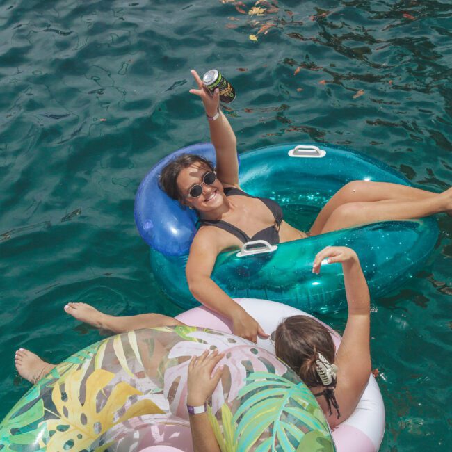 Two women relax on inflatable pool floats in clear blue water. One woman, wearing sunglasses and a black bikini, smiles and raises a can in one hand. The other lounges nearby on a pink and green float.
