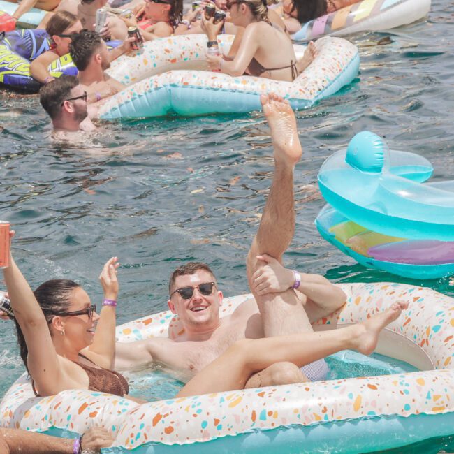 People relaxing in colorful inflatable pool floats on water at a lively outdoor party. A smiling man and woman enjoy drinks and pose playfully in a sprinkle-patterned float. Others float and socialize in the background.