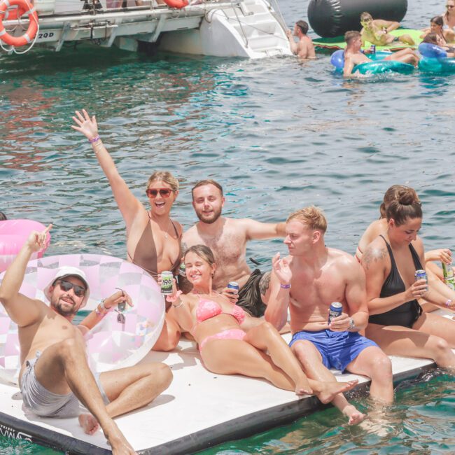 A group of people in swimsuits sit and pose on a floating platform in the water, smiling and holding drinks. Others float nearby on inflatable pool toys, with a docked boat in the background. The mood is joyful and festive.