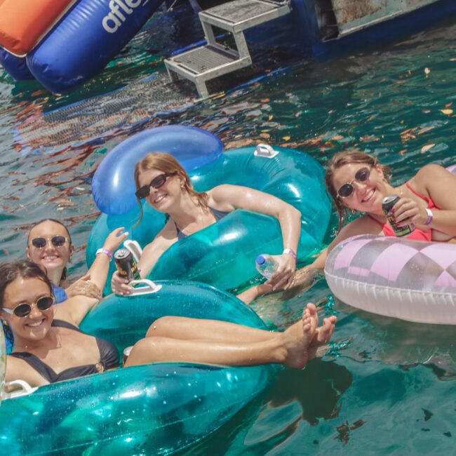 Four women in swimsuits and sunglasses smile while relaxing on colorful inflatable floats in the water, holding drinks. Boats and water toys are visible in the background. The scene is bright and cheerful.