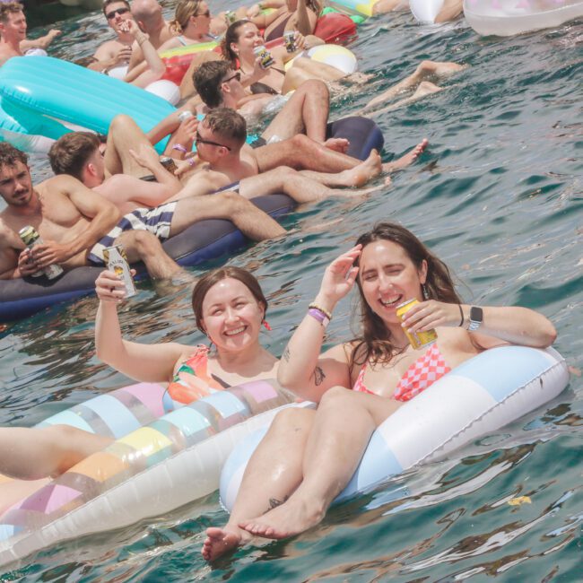 A group of young adults relax on colorful pool floats in a sunny, outdoor body of water. Two women in the foreground smile and hold drinks, enjoying the lively atmosphere with others in the background.