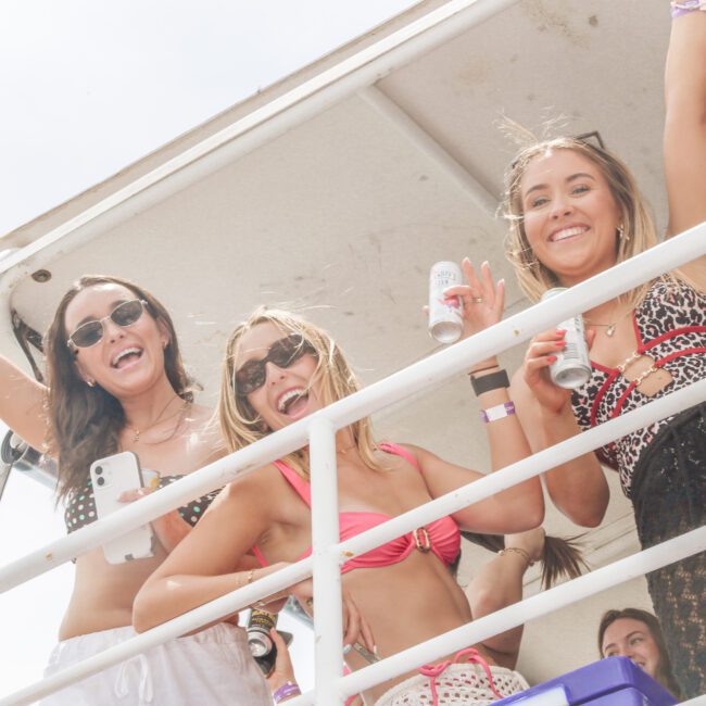 Four women in swimsuits and sunglasses smile and pose with drinks on an outdoor balcony, enjoying a sunny day. One holds up her arm while others lean on the railing, looking cheerful and relaxed.