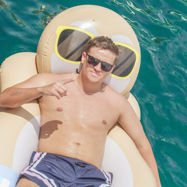 A man wearing sunglasses and swim trunks relaxes on a large inflatable float shaped like an emoji with sunglasses while floating on clear blue water, smiling and pointing at the camera.