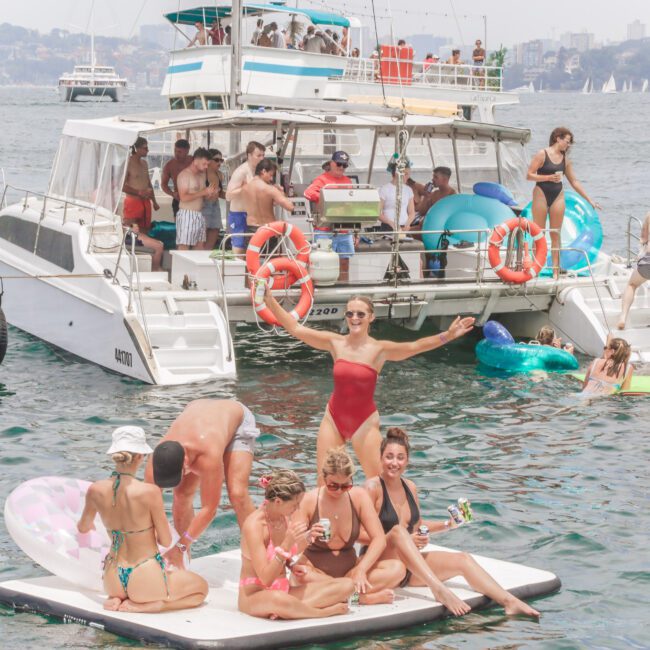 A group of people relax on floats and paddleboards near a crowded party boat on the water. Some are holding drinks, while others stand or pose, enjoying a sunny day with a city skyline in the background.